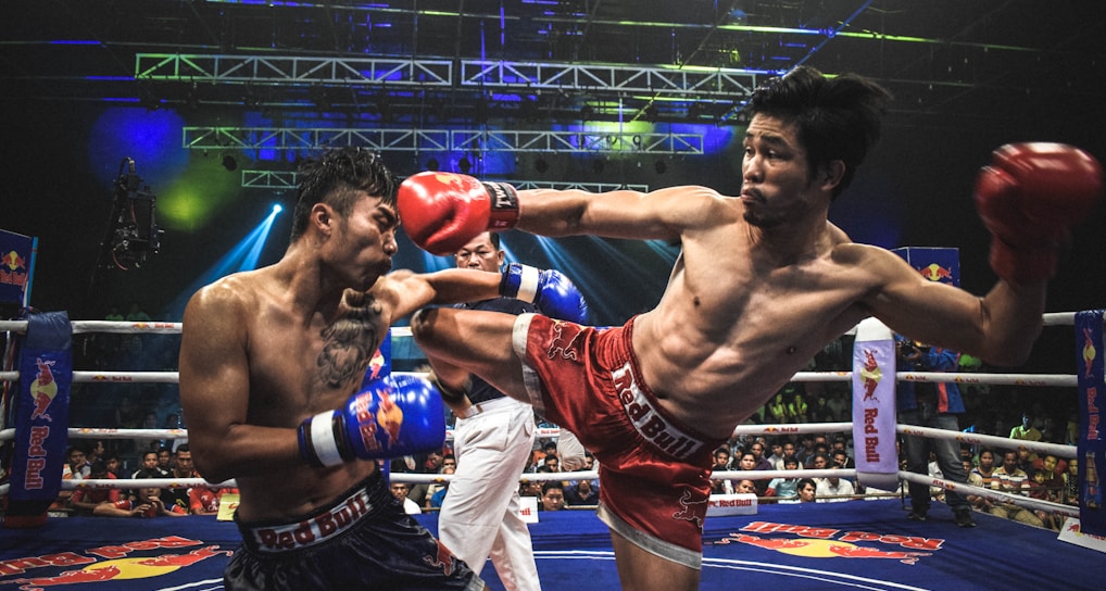 Two athletes are engaged in a dynamic kickboxing match inside a ring, surrounded by a cheering audience. One fighter, wearing red gloves and shorts, executes a high kick towards his opponent, who attempts to block the attack with his arm. The background features colorful lighting and promotional banners.