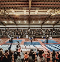 A large indoor arena with a high ceiling, filled with an audience seated on bleachers. In the center, two fighters are engaged in a martial arts competition on blue and red mats. The scene is captured through a mesh fence, adding a textured foreground.
