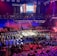 An indoor arena is filled with people watching a wrestling match. The ring is centrally located, and surrounded by audience members sitting in tiered seating. The arena is illuminated with red and blue lights, and a large overhead screen displays 'Sunday Stunner.' Spectators are engrossed in the match taking place in the well-lit ring.