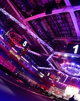 An indoor arena with vibrant purple and blue lighting, featuring large numbered screens suspended from the ceiling. The venue has a high ceiling with intricate metal truss structures and hanging lights, creating a futuristic and energetic atmosphere.