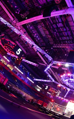 An indoor arena with vibrant purple and blue lighting, featuring large numbered screens suspended from the ceiling. The venue has a high ceiling with intricate metal truss structures and hanging lights, creating a futuristic and energetic atmosphere.